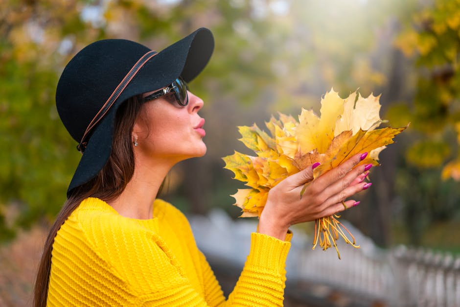 Woman in yellow sweater and hat holding autumn leaves outdoors.