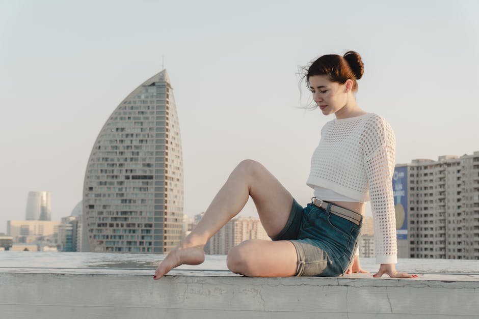 A young woman sits on a rooftop with cityscape in the background under a bright sky.