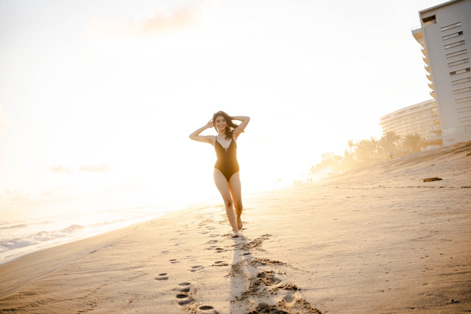 A woman in a swimsuit walks on a sunlit beach with footprints and a modern skyline.