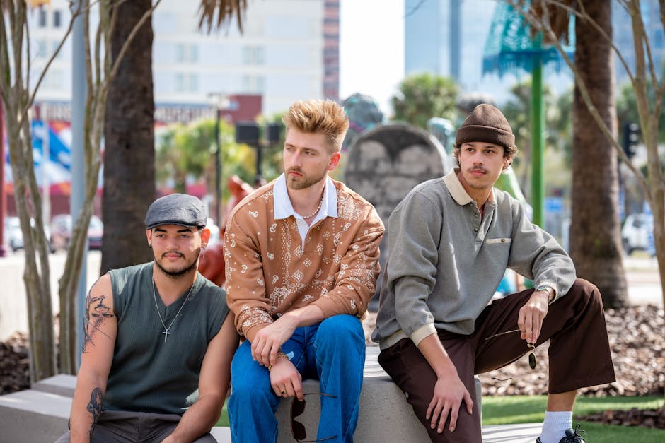Three fashionable young men sitting outside in a city park, showcasing modern street style and confidence.